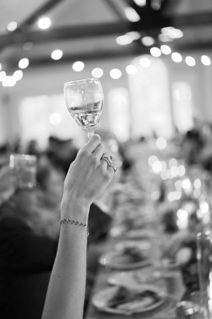 A person holds up a wine glass in a toast at a long, candlelit table during a festive gathering. The background is softly blurred, highlighting the celebratory atmosphere.