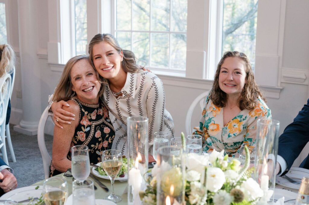 Three women sit at a table decorated with white flowers and glasses, smiling at the camera; one young woman hugs an older woman while another woman sits beside them, all in a bright room with large windows.