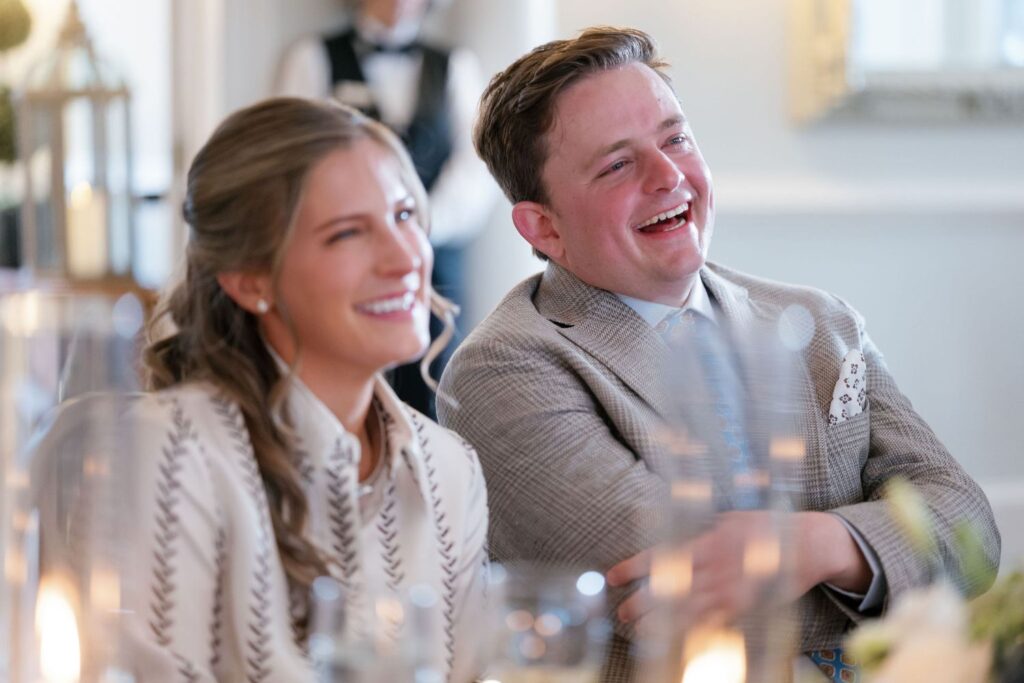 A smiling woman and man dressed formally sit together at a table, enjoying an event. The background is softly blurred, with candles and another person in the distance. Both appear happy and engaged.