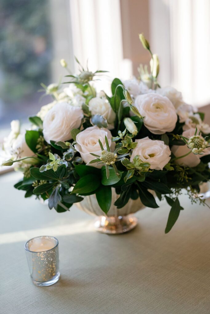 A silver vase filled with white roses and greenery sits on a light green tablecloth next to a small glass candle holder with a lit candle. Sunlight shines through windows in the background.