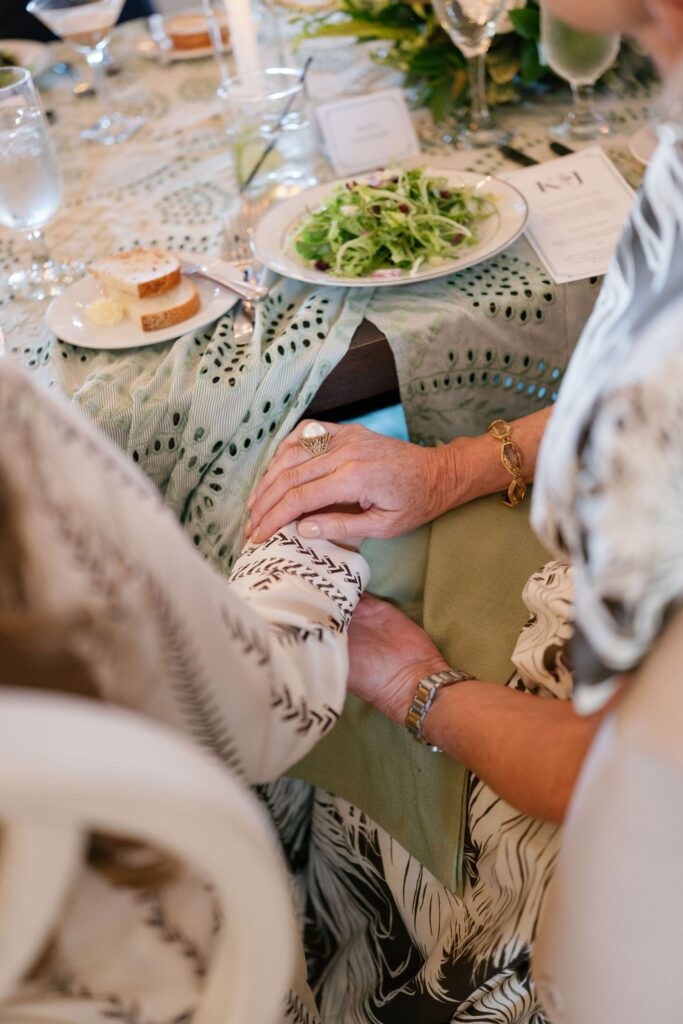 Two women seated at a table hold hands over a green napkin. The table is set with a salad, bread, glasses, and a printed menu. The scene conveys warmth and connection at a formal meal.