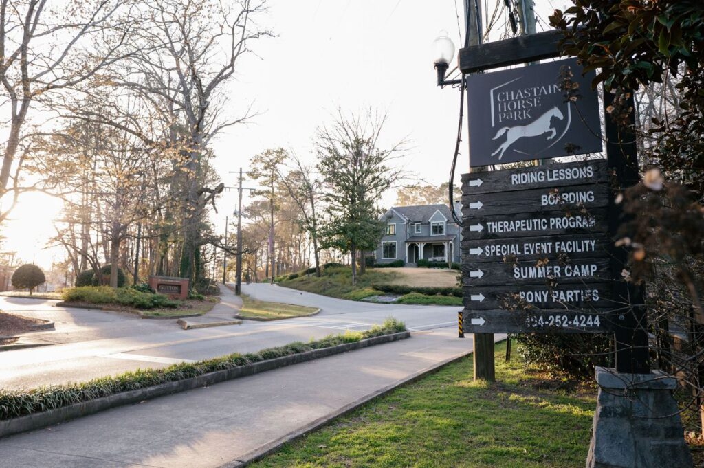 A wooden sign for Chastain Horse Park lists services like riding lessons and summer camp. A house and trees are in the background, with sunlight shining through branches along a quiet road.