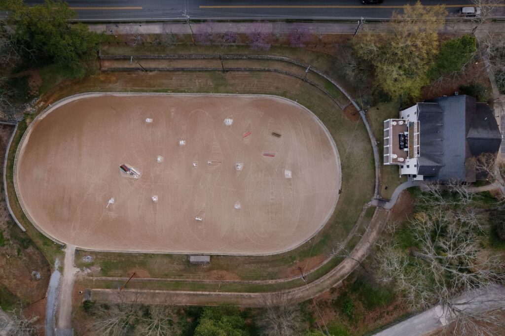Aerial view of a horse riding arena with jumps set up, next to a large house. The arena is oval-shaped, surrounded by trees and paths, with a road running along the top of the image.