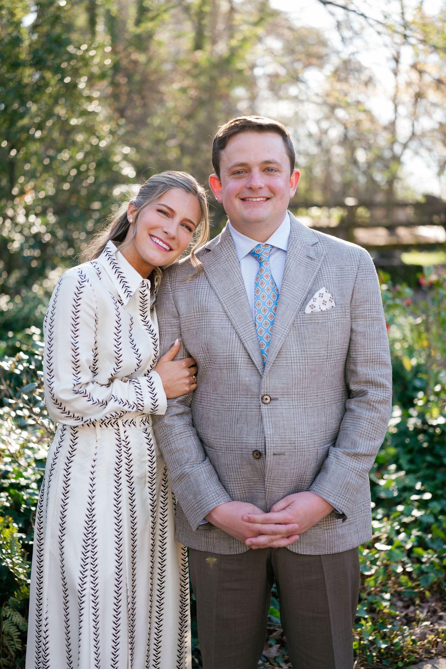 A woman in a patterned white dress leans affectionately on a smiling man in a light gray suit and blue tie. They stand outdoors in a sunlit garden with green foliage in the background.