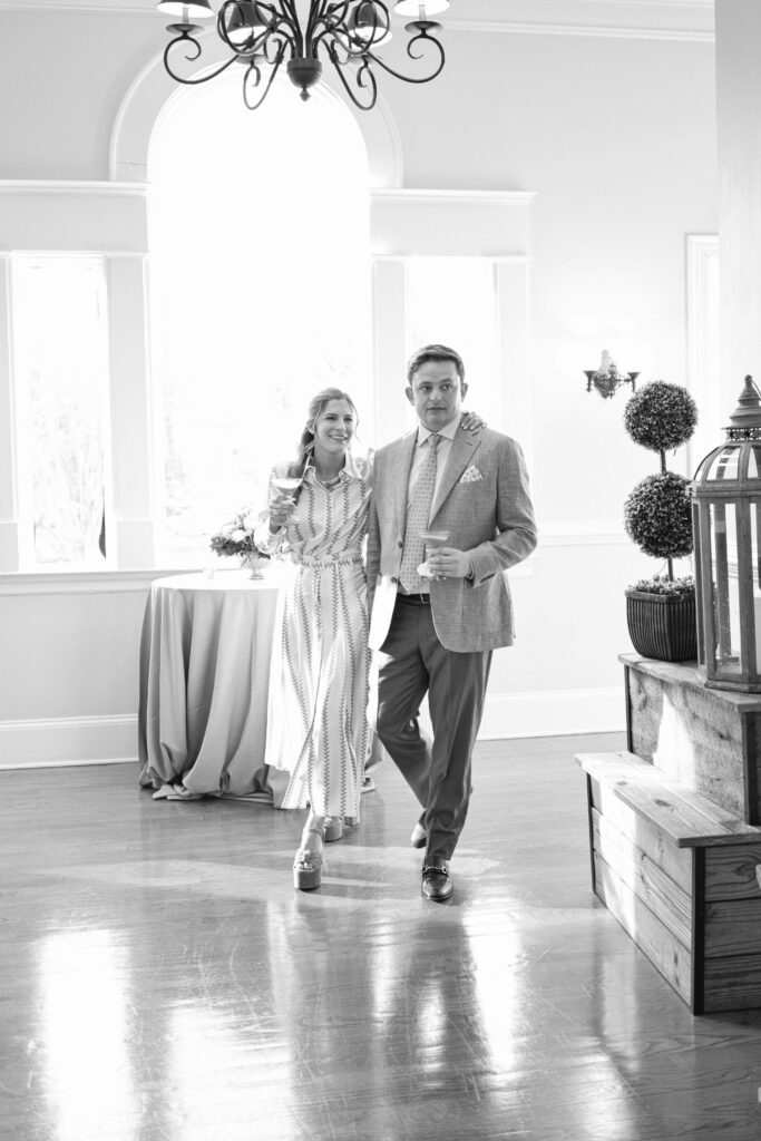 A woman in a long dress and a man in a suit walk arm-in-arm across a bright room with large windows and elegant decor, including potted plants and stacked wooden crates.