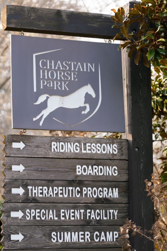 A wooden sign for Chastain Horse Park with a horse logo and arrows pointing to riding lessons, boarding, therapeutic program, special event facility, and summer camp.
