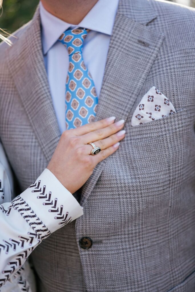 A person in a patterned suit and tie stands as another person gently rests a hand with a ring on their chest, near the suits pocket square.