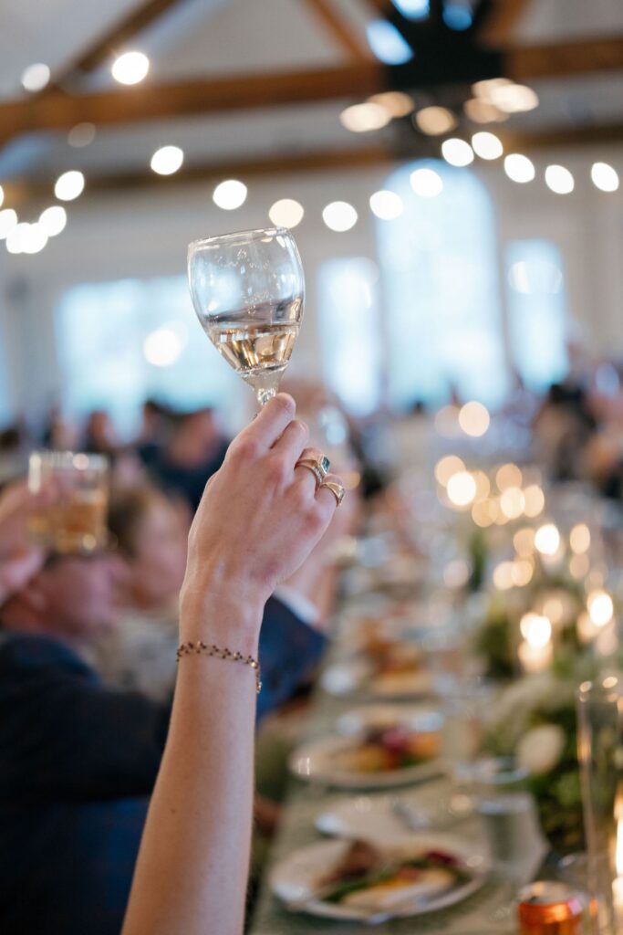 A hand holding up a glass of white wine in a toast at a festive, candle-lit dinner with blurred guests and plates visible in the background.