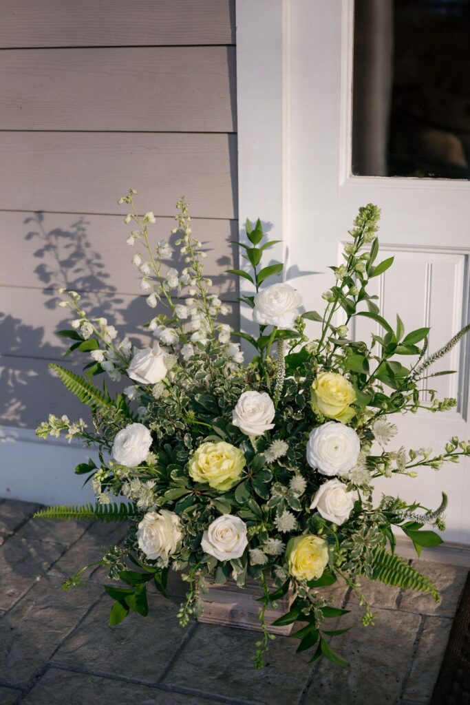 A floral arrangement with white and pale yellow roses, green ferns, and assorted greenery sits on a stone patio in front of a light gray wall and white door.