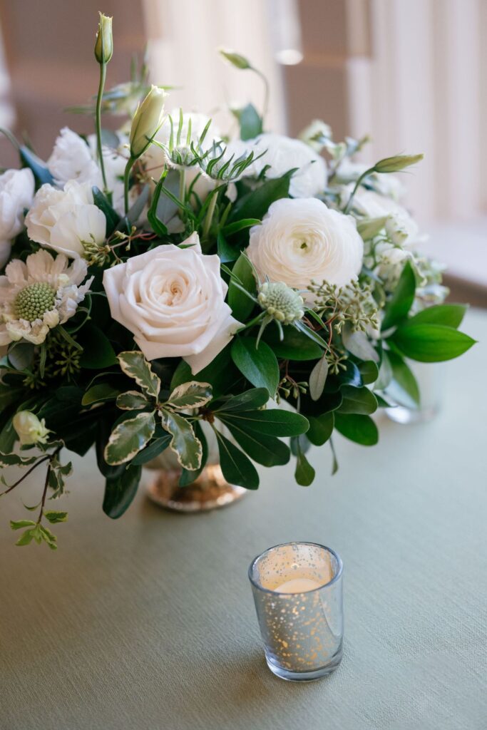 A small glass votive candle sits in front of a floral arrangement with white roses, ranunculus, greenery, and other white flowers on a light green tablecloth.