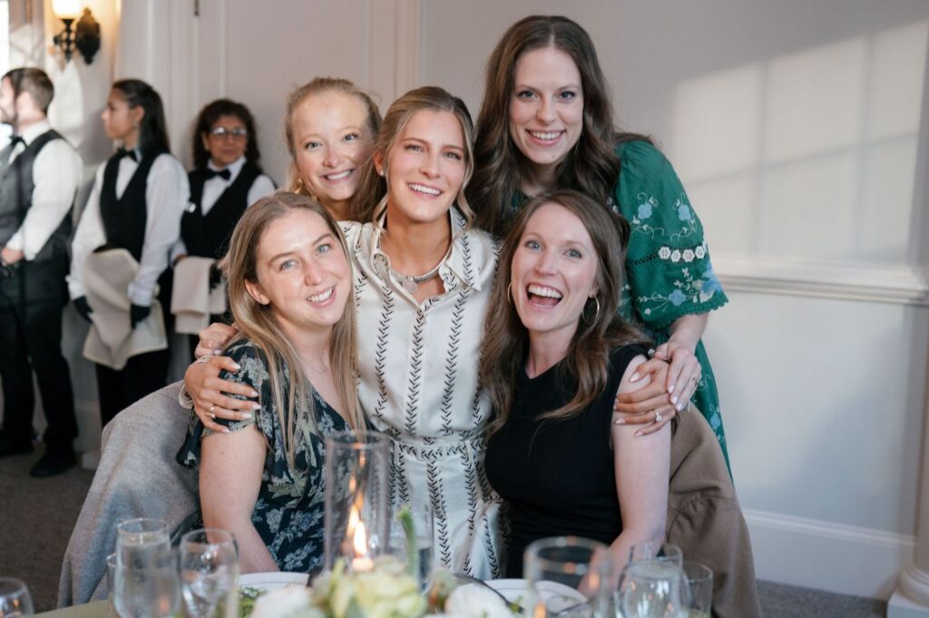 Five women smiling and posing together at a table during an indoor event; some dressed in casual and some in semi-formal attire. In the background, servers in black and white uniforms stand near a wall.