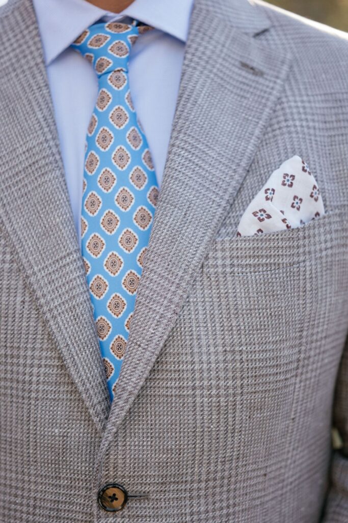 A close-up of a man wearing a light gray plaid suit, light blue dress shirt, blue and beige patterned tie, and a white pocket square with a brown design.