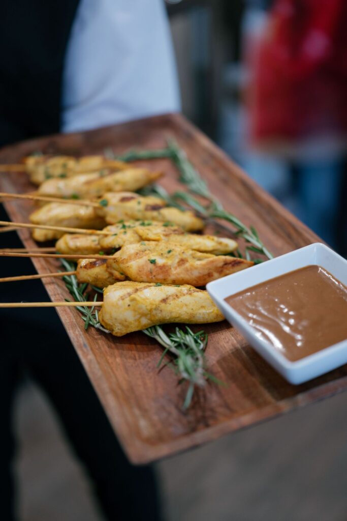 Grilled chicken skewers served on a wooden platter with fresh rosemary and a small square dish of brown dipping sauce.