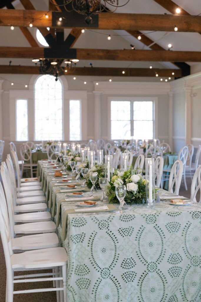 Elegant dining setup with white chairs around a long table covered in a light green patterned tablecloth, floral centerpieces, glassware, and tall candles in a bright room with large windows and exposed wooden beams.