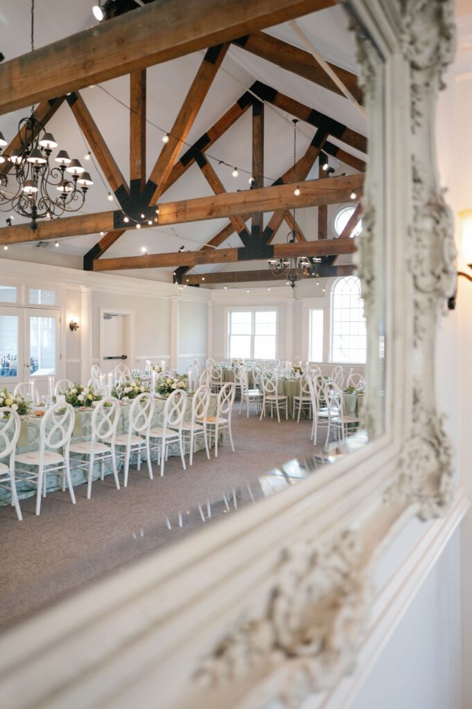 Elegant reception hall reflected in a decorative mirror, featuring long tables with white chairs, floral centerpieces, high wooden beam ceiling, chandeliers, and large windows letting in natural light.