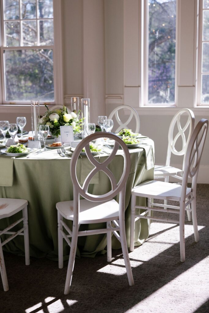 A round table with a light green tablecloth is set for a meal, featuring elegant white chairs, glassware, plates with salad, and a floral centerpiece. Sunlight streams through large windows, brightening the room.