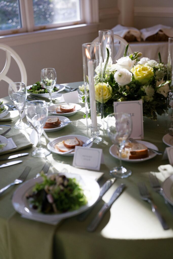 A beautifully set dining table with plates of salad, bread, wine glasses, candles, and a floral centerpiece of white and yellow flowers, illuminated by natural light from nearby windows.