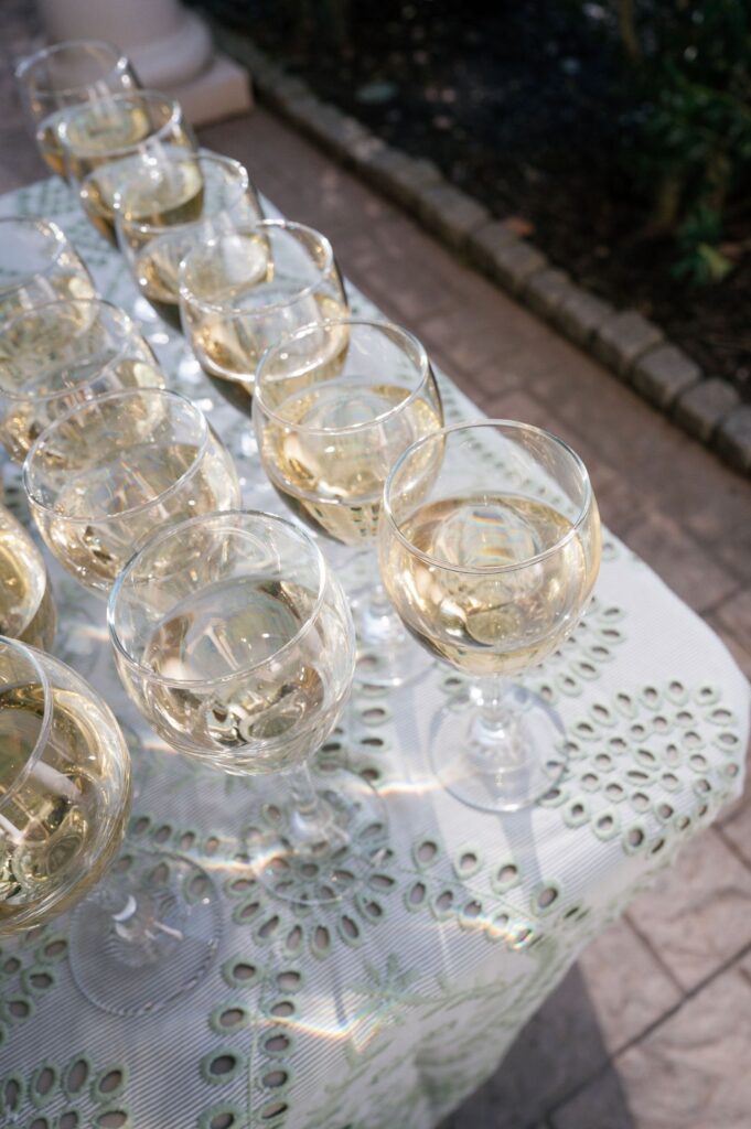 Twelve glasses of white wine arranged on a patterned white tablecloth outdoors, with sunlight creating reflections on the table.