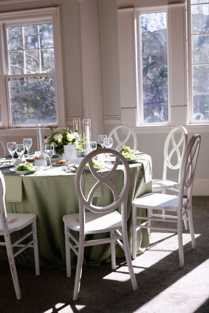 A round table covered with a sage green tablecloth is set with plates, glasses, and green salads. White chairs surround the table, and sunlight streams in through tall windows in the background.