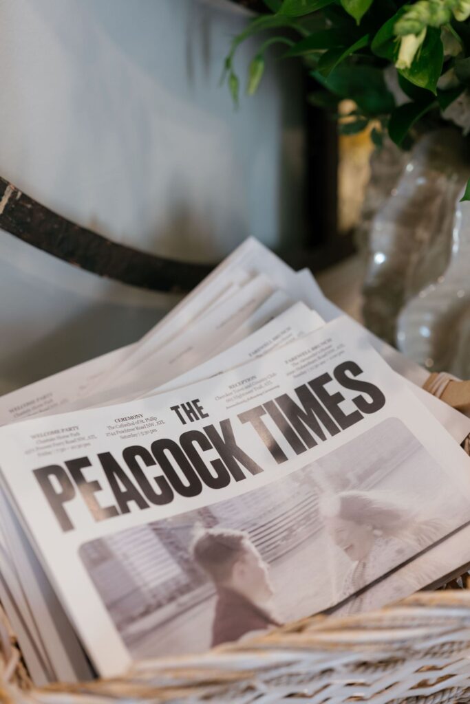 A stack of newspapers titled The Peacock Times sits in a wicker basket, with blurred images of two people on the front page and some green foliage visible in the background.
