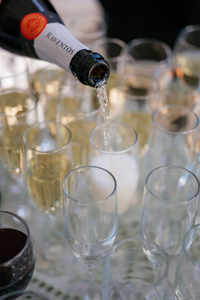 A close-up of sparkling wine being poured from a Raventós bottle into a clear champagne glass, surrounded by other filled and empty glasses on a table.