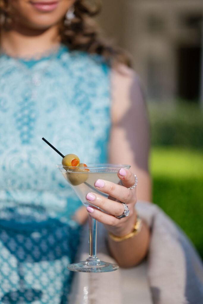A woman wearing a sleeveless teal lace dress holds a martini glass with olives on a cocktail pick. She has light pink nails, rings, and a gold bracelet, and is standing outdoors with greenery in the background.
