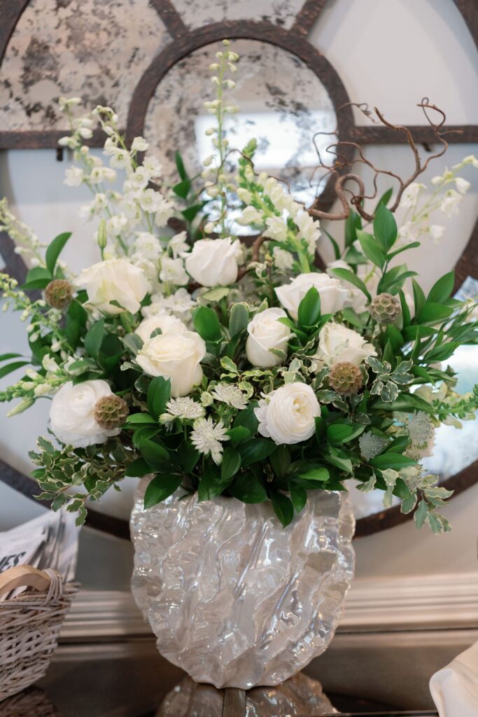 A decorative arrangement of white flowers and greenery in a textured, translucent vase sits on a table, with a large rustic mirror in the background.
