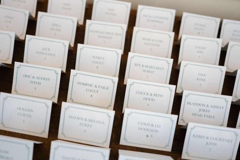 A close-up of several name cards arranged in rows on a wooden surface, likely serving as seating assignments for an event such as a wedding or formal gathering. Each card lists guest names and table numbers.