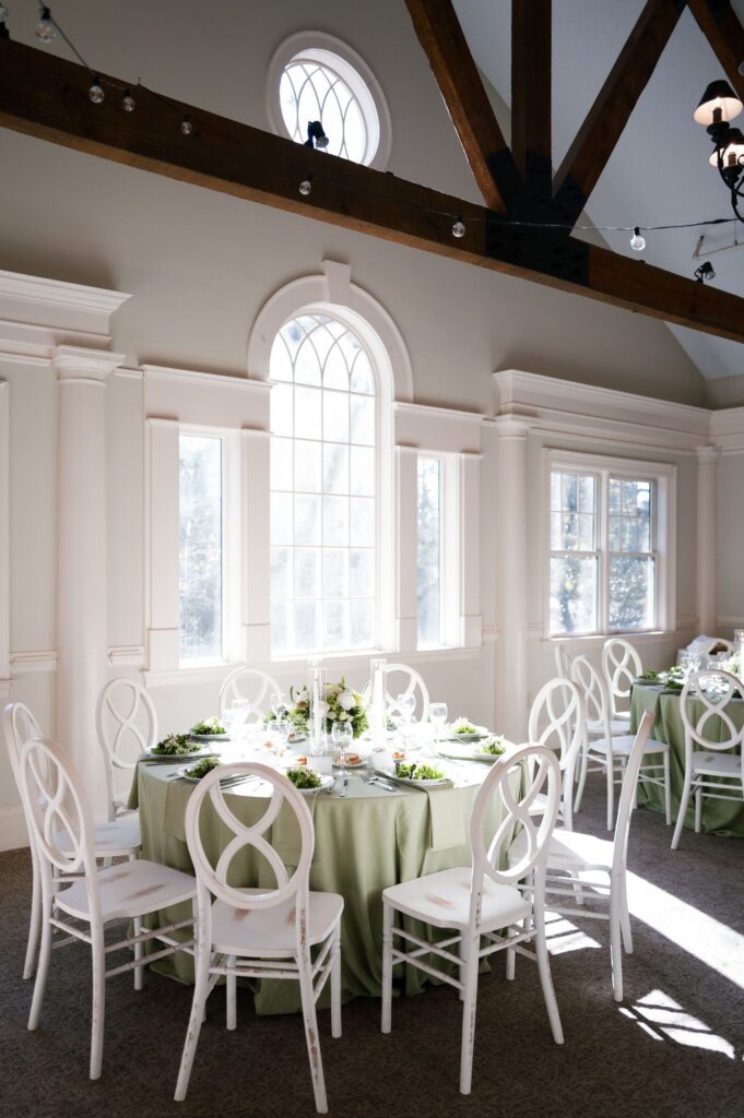 A round table with a pale green tablecloth is set for a formal meal, surrounded by white chairs in a bright room with large arched windows, white walls, and exposed wooden beams. Sunlight streams through the windows.