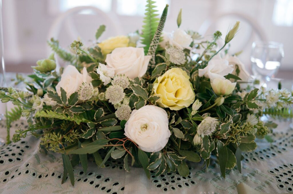 A floral centerpiece with white and pale yellow roses, green leaves, and ferns arranged on a lace tablecloth.