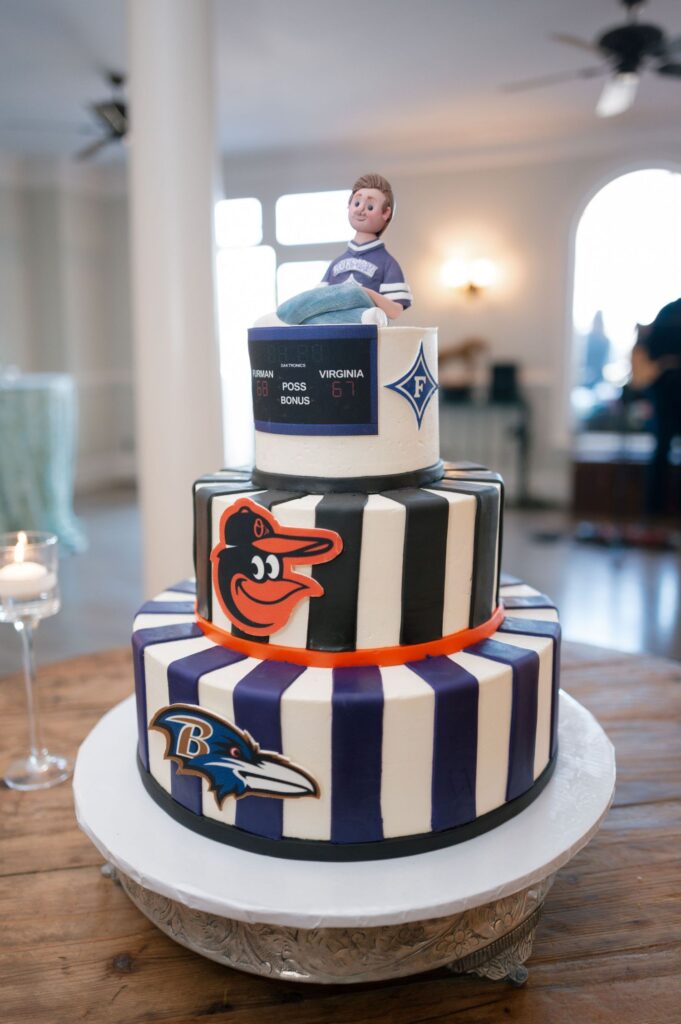 Three-tier cake decorated with black and white stripes, featuring Baltimore Orioles and Ravens logos. The top tier has a small figure of a boy in a sports jersey and scoreboard details. The cake is on a table indoors.