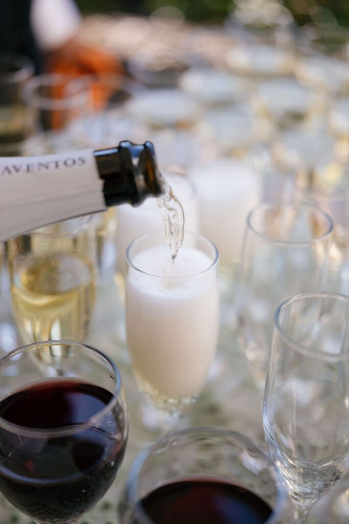 A close-up of sparkling wine being poured into a flute glass, creating bubbles, surrounded by various glasses filled with wine and champagne on a table.