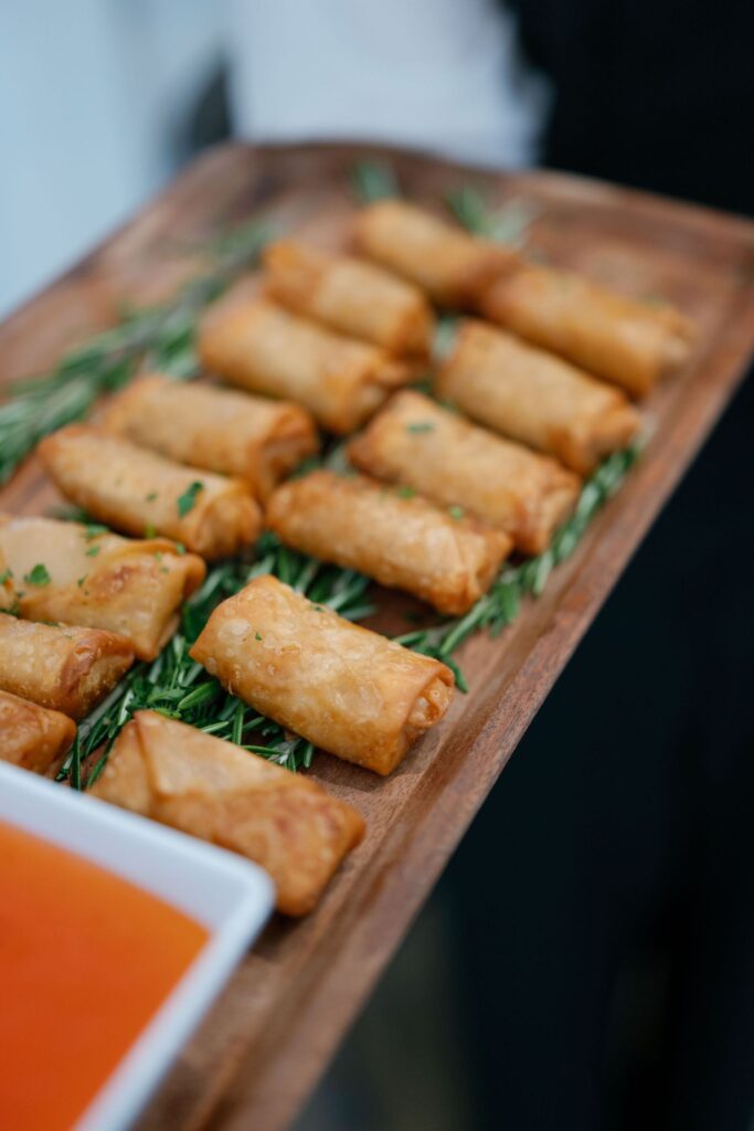 A wooden tray holds several crispy spring rolls on a bed of fresh herbs, with a bowl of orange dipping sauce in the corner.