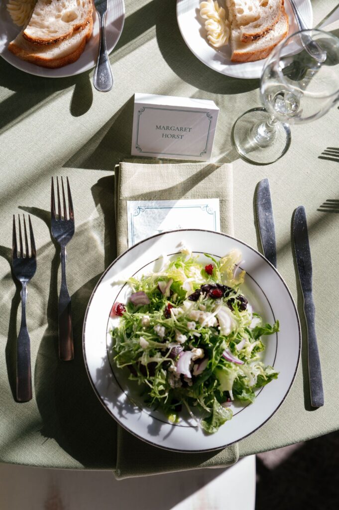 A plate of mixed green salad with red onions and cranberries sits on a table with a place card reading Margaret Horst, next to silverware, a wine glass, and a plate of bread with butter.