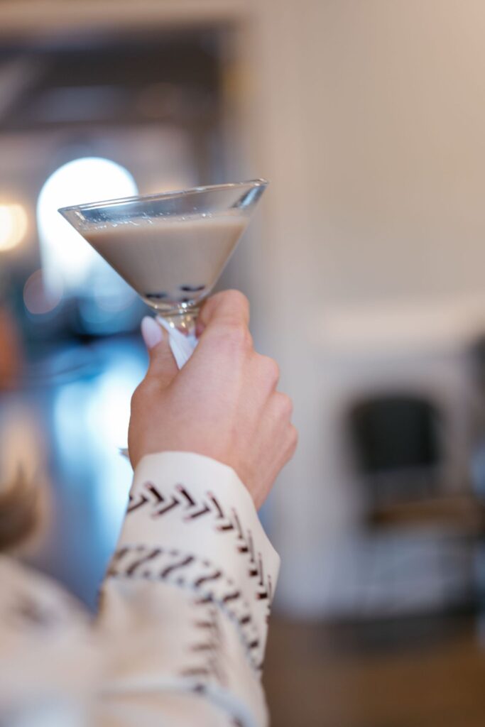 A hand holding a martini glass filled with a creamy, light-colored cocktail. The person wears a light-colored sleeve with a dark, stitched pattern. The background is softly blurred.