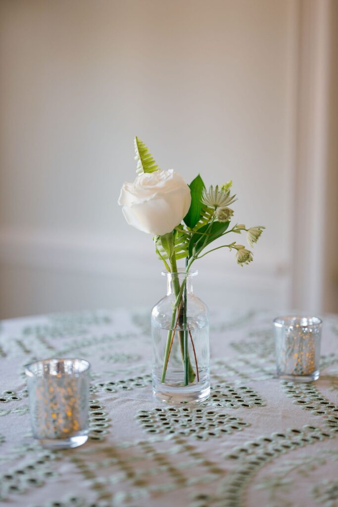 A small glass vase with a single white rose and green leaves sits on a patterned tablecloth, flanked by two silver candle holders. The background is softly blurred.