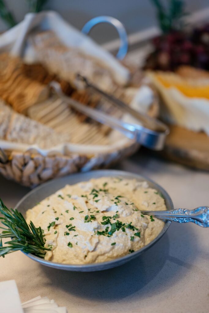 A bowl of creamy dip garnished with chopped herbs and a sprig of rosemary sits on a table. In the background, a basket with assorted crackers and tongs is slightly out of focus.