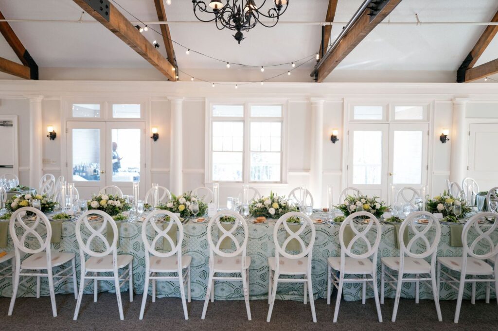 A long banquet table set for a formal event, decorated with white flowers, greenery, glassware, and white chairs, beneath a chandelier and exposed wooden beams in a bright, elegant room.