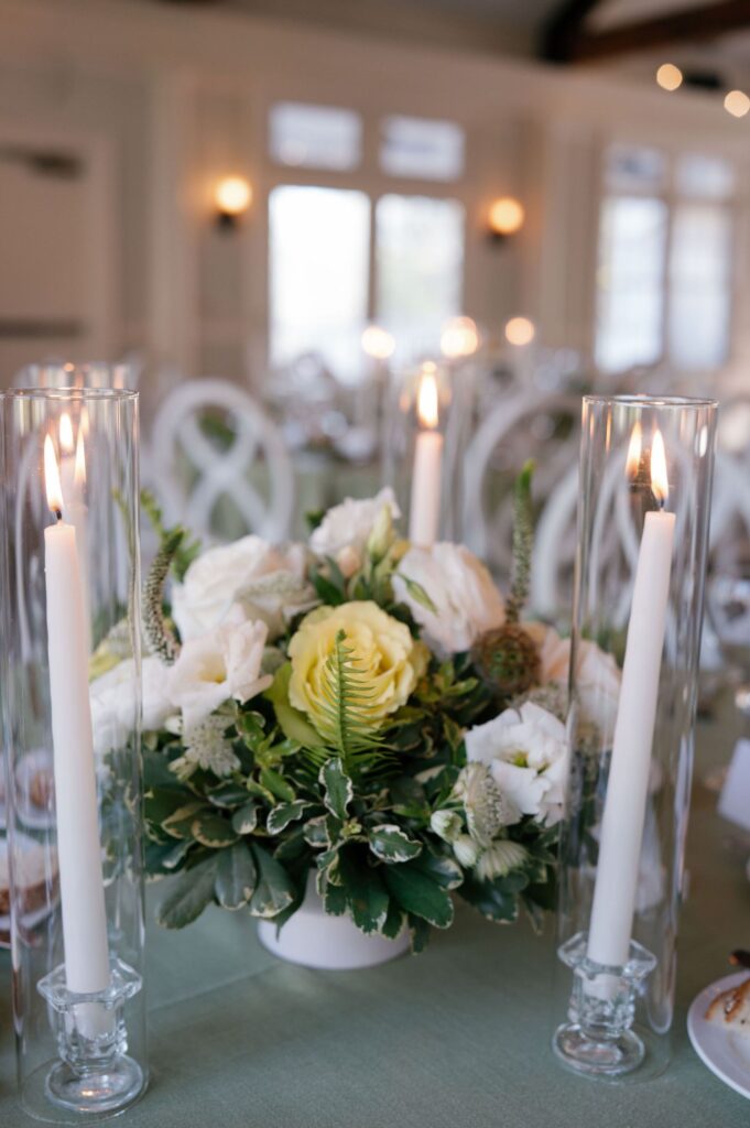 A floral centerpiece with white and pale yellow flowers and greenery sits on a table, flanked by two tall glass candle holders with lit white candles in a softly lit, elegant room.