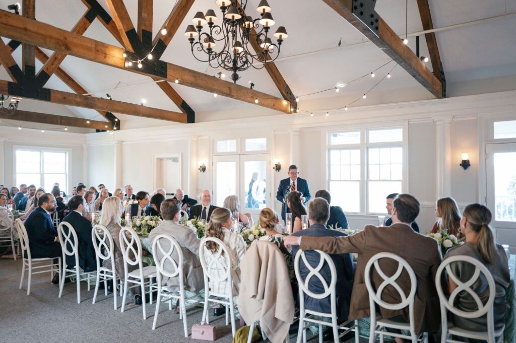 A man stands and speaks at the head of a long, elegantly set table during a formal event in a bright room with exposed wooden beams and a chandelier. Guests sit and listen attentively.