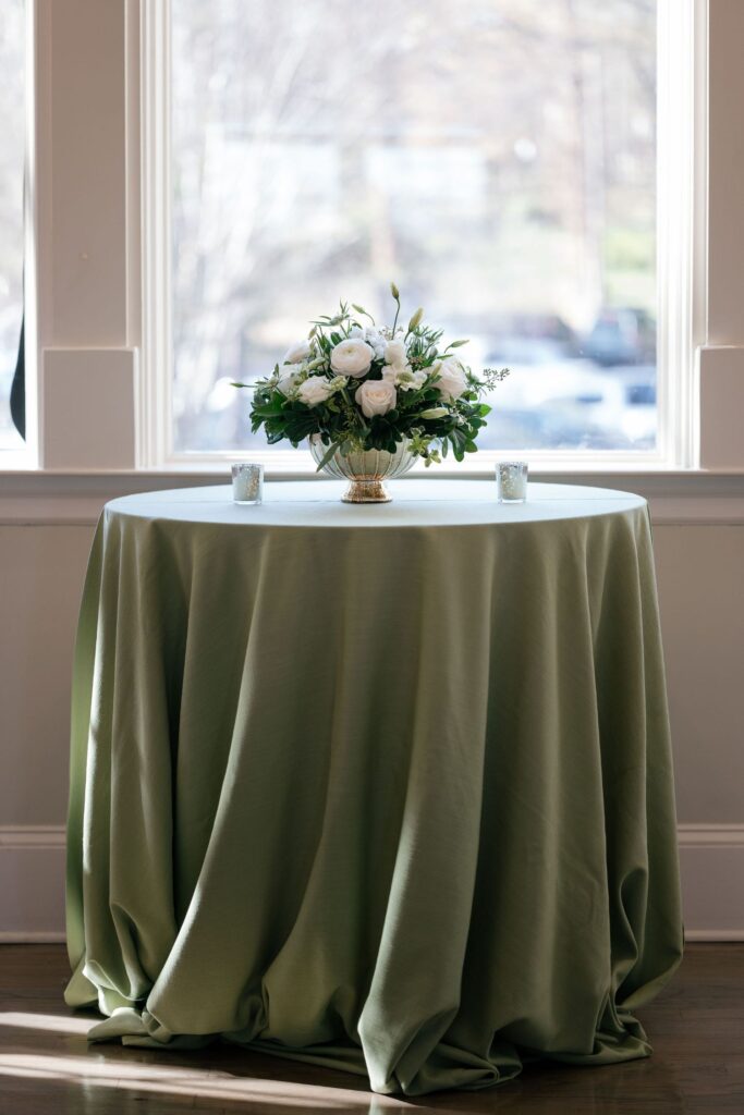 A round table covered with a sage green tablecloth is set in front of a large window. A floral arrangement of white flowers and greenery sits at the center, flanked by two small votive candles.