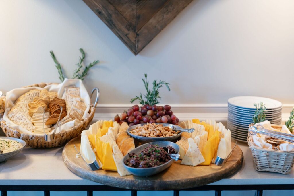 A wooden platter with assorted cheeses, grapes, bread, nuts, and dips, surrounded by baskets of crackers and a stack of white plates, arranged on a countertop for a gathering.