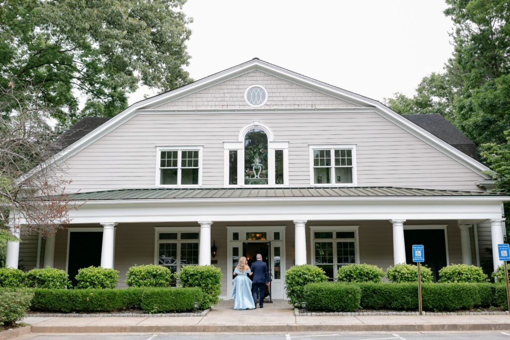 A large, light gray house with white pillars and green shrubs out front hosts a progressive cocktail reception in Atlanta. Two people, one in a light blue dress and the other in dark clothing, stand near the entrance amid tall surrounding trees.