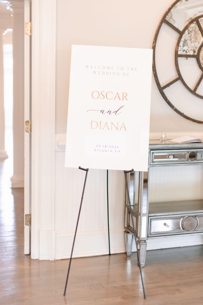 A white wedding welcome sign on an easel reads “Welcome to the wedding of Oscar and Diana, 05/18/2024, Atlanta, GA” in gold and blush lettering. Perfect for a progressive cocktail reception Atlanta guests will remember, it stands in a bright, elegant room.