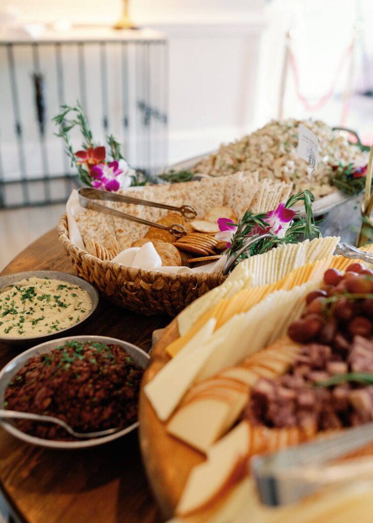 A wooden table set for a progressive cocktail reception Atlanta, featuring a variety of sliced cheeses, crackers, bread, dips, and garnishes, beautifully arranged with fresh herbs and edible flowers for an inviting buffet or party.
