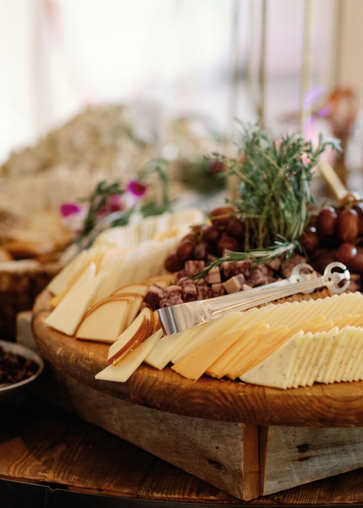 A wooden platter displays an assortment of sliced cheeses, grapes, and herbs, with metal tongs resting on top—perfect for a progressive cocktail reception in Atlanta; the background is softly blurred.