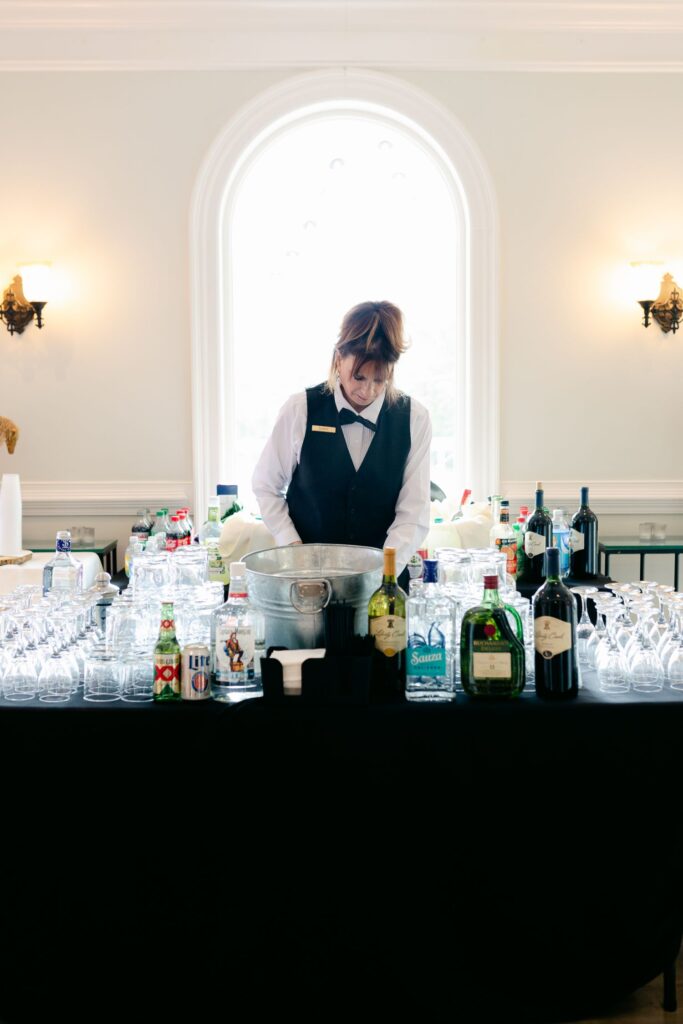 A bartender in a black vest stands behind a table with bottles of liquor, mixers, and glasses, preparing drinks for a progressive cocktail reception in Atlanta, set in a bright room with a large arched window and elegant wall sconces.