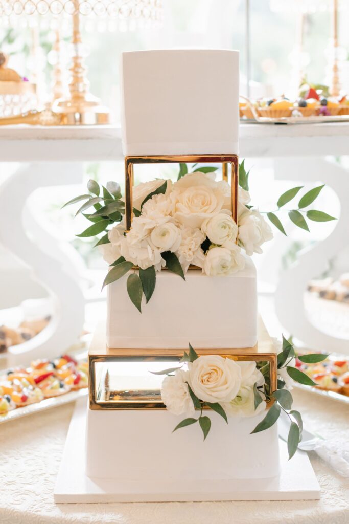 A three-tiered white wedding cake with gold square frames between the tiers, decorated with white roses and green leaves, is displayed on a table—perfect for a progressive cocktail reception in Atlanta, with assorted desserts in the background.