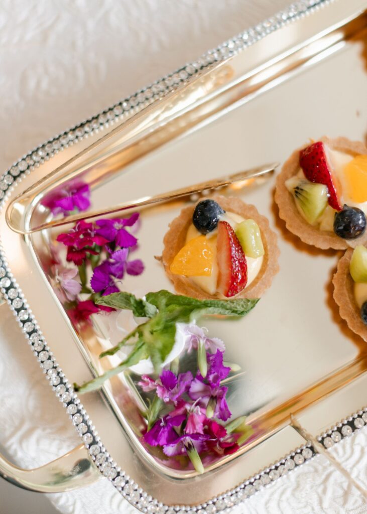 A silver tray with beaded edges holds small fruit tarts topped with strawberries, kiwi, orange, and blueberries—perfect for a progressive cocktail reception in Atlanta—alongside a garnish of purple flowers and green leaves.