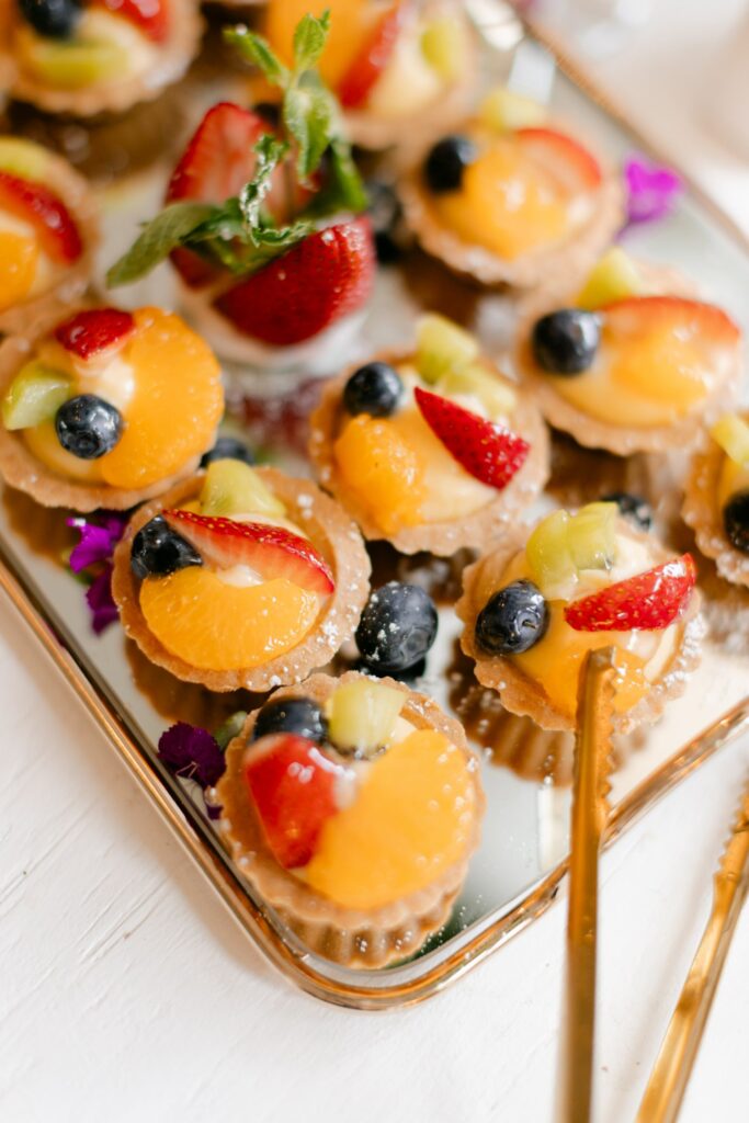 A tray of mini fruit tarts topped with glazed strawberries, blueberries, kiwi, and orange segments, adorned with mint leaves and served with gold utensils—perfect for a progressive cocktail reception in Atlanta.
